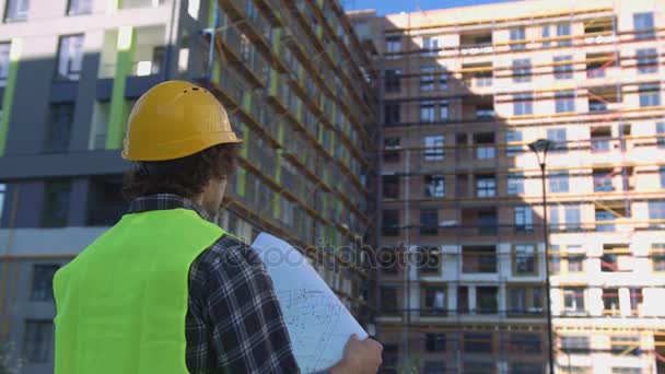 Back view of builder in protected helmet and vest looking drawing on unfinished construction background. - Felvétel, videó
