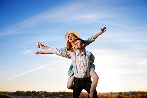 Young guy piggybacking cheerful girlfriend like airplane on the background of sky - Photo, Image