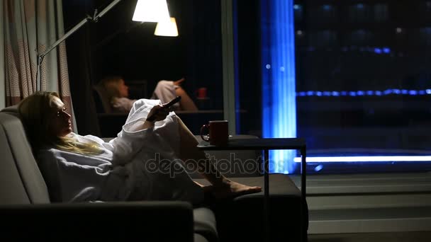 Een vrouw is liggend op een bank met een mok in haar handen in de buurt van het venster met een nacht uitzicht over de stad. Tv kijken - Video