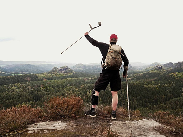 Hombre feliz excursionista sosteniendo muleta medicina por encima de la cabeza, rodilla lesionada fijada en la rodilla característica
.  - Foto, imagen