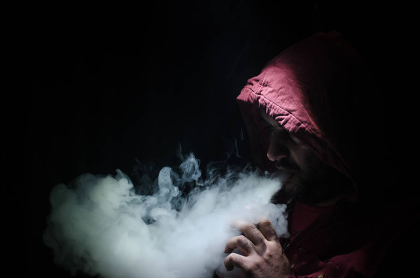 Vaping man holding a mod. A cloud of vapor. Black background. Vaping an electronic cigarette with a lot of smoke - Фото, зображення