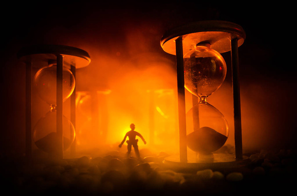 Концепция времени Силуэт человека, стоящего между песочными часами с дымом и огнями на темном фоне
. - Фото, изображение