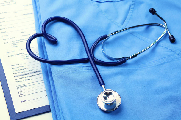 Medische stethoscoop twisted in hart vorm liggen op lijst patiënt medische geschiedenis en blauwe arts uniforme closeup - Foto, afbeelding