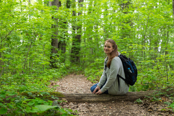 Tourist girl with backpack is sitting on the tree bark in the green forest - Fotó, kép