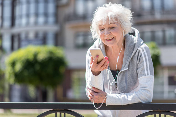 Cheerful old sporty woman listening to music via headphones - Foto, Imagem