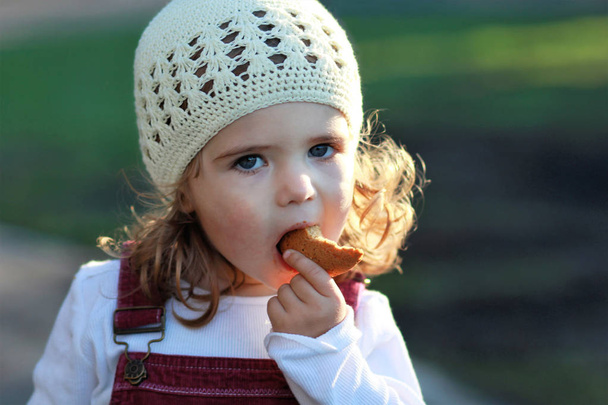 Close-up van portret van leuk één-jarige meisje in een witte gebreide muts cookie eten op een wandeling in het park - Foto, afbeelding