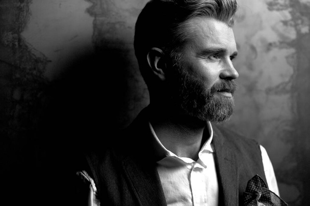 Great portrait of handsome serious charismatic man with beard, in white shirt and black classic waistcoat, posing in studio on dark grey background, in black and white - Фото, изображение