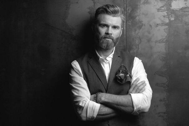 Handsome serious charismatic man with beard, in white shirt and black classic waistcoat, thinking and posing in studio on dark grey background, in black and white - Fotografie, Obrázek