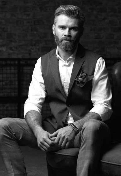Handsome serious charismatic man with beard, in white shirt and black classic waistcoat, siting in studio on dark grey background, in black and white - Valokuva, kuva
