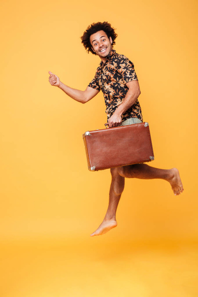 Cheerful african man jumping holding suitcase and showing thumbs up. - Foto, afbeelding
