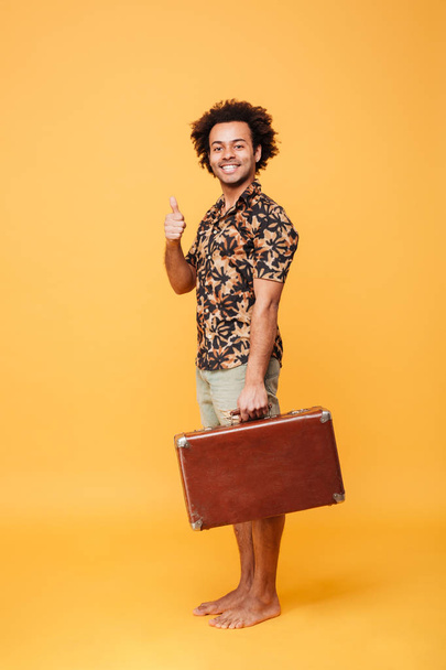 Happy young african man holding suitcase showing thumbs up. - Фото, изображение