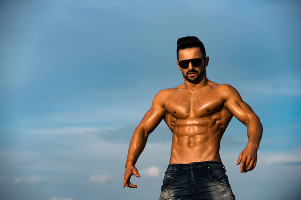 homem sexy com corpo muscular em óculos de sol no céu azul
 - Foto, Imagem