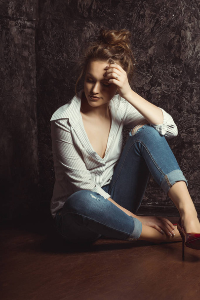 Τρυφερή νεαρή γυναίκα στο πουκάμισο και τζιν που κάθεται στο πάτωμα - Φωτογραφία, εικόνα