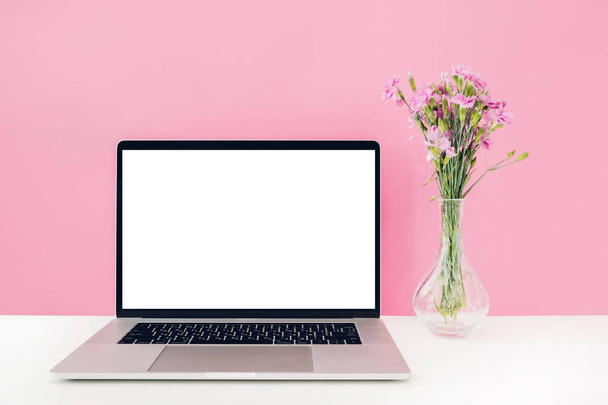 Laptop with white blank screen and flowers in vase on table on pink background. mock up - Fotoğraf, Görsel
