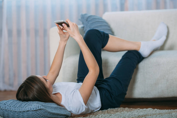 Mladá žena kontrolu její chytrý telefon leží na koberci - Fotografie, Obrázek