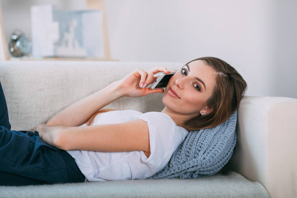 Young woman calling by her smart phone lying on sofa - Foto, Bild