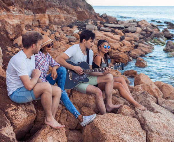 friends sitting on stones on beach. man is playing guitar. - Fotoğraf, Görsel