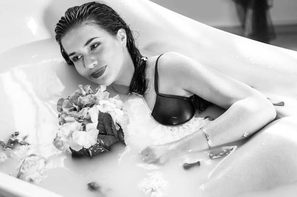 Sexy brunette vrouw ontspannen in hete melk bad met bloemen - Foto, afbeelding