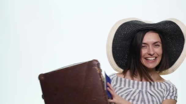 Portrait of trendy young female in wicker hat standing with suitcase and holding passport with tickets over white background - Imágenes, Vídeo