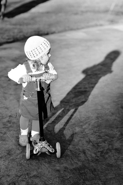 Black and white portrait of a stylish one year old girl riding a scooter and her shadow on a lovely summer day - Photo, Image