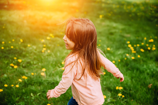 Petite fille courant le long de la pelouse verte avec des pissenlits jaunes, vue de dos. Image de fond, concept sur le thème du bonheur, de l'enfance et de l'insouciance
. - Photo, image