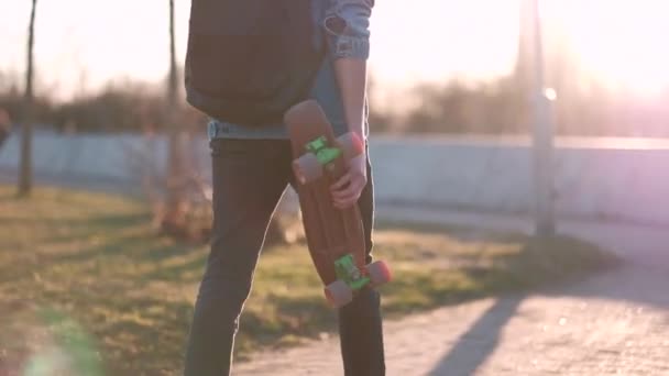 若い男が手にスケートボードで歩く,日没時,デニムジャケットとブラックジーンズで - 映像、動画