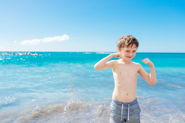 Αυτοπεποίθηση μικρό αγόρι που δείχνει τους μυς του παίζοντας στην παραλία - Φωτογραφία, εικόνα