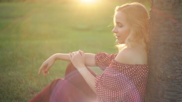 Młoda piękna kobieta siedzi pod drzewem na trawie w zachód słońca - Materiał filmowy, wideo