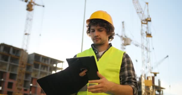 Concentrated young builder in yellow helmet and green vest typing on tablet for work on unfinished construction background. - Materiaali, video