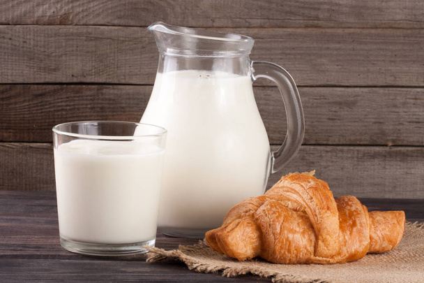 kruik en glas melk met croissant op een houten achtergrond - Foto, afbeelding