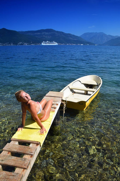 jolie jeune blonde en bikini souriant vers la caméra tout en bronzant au bord de l'eau, bateau de croisière en arrière-plan
 - Photo, image