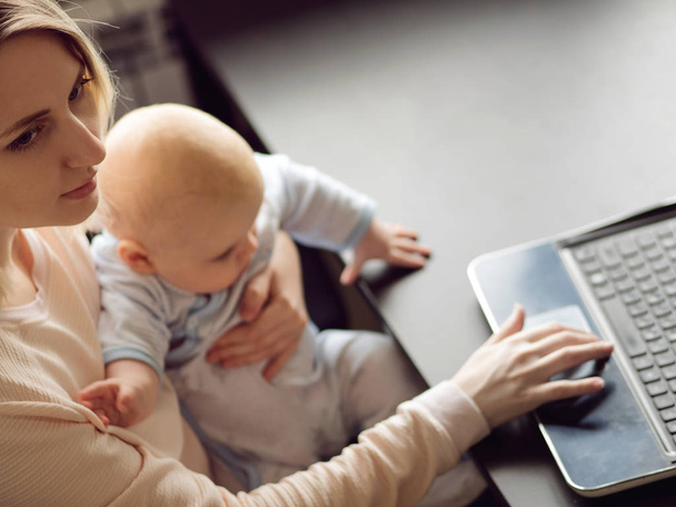 Young beautiful blonde woman, mother in home office with computer and her baby. Work and child care. - Foto, afbeelding