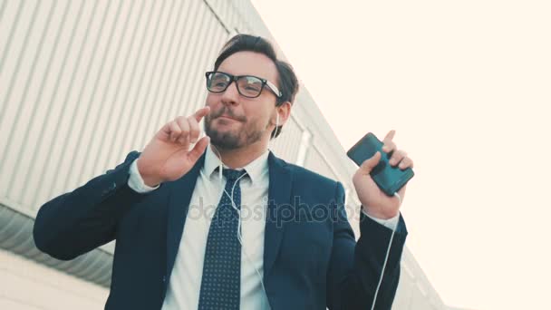 Happy handsome businessman listening to music in headphones on smartphone, walking outside the office and funny dancing to the rythm. Funny dancing moves. Close up - Séquence, vidéo