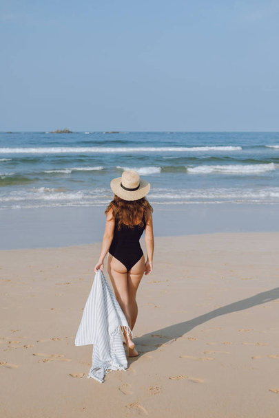 Girl in a black swimsuit and hat walking on the beach - Foto, Imagen