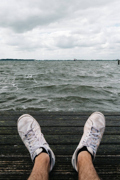 Homme assis sur la jetée avec des baskets au premier plan et le paysage marin sur
  - Photo, image