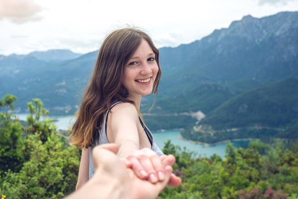 Pójdź za mną, atrakcyjny brunetka dziewczynka trzymając się za ręce z potencjalnymi klientami w górskiej dolinie z rzeki - Zdjęcie, obraz