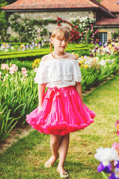 Outdoor portrait of adorable little girl playing in a beautiful flower park on a nice summer day. Fashion kid girl wearing white blouse and bright pink tutu skirt - Photo, Image