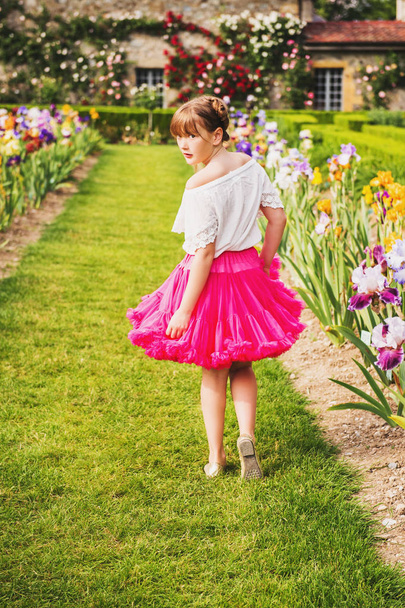 Venkovní portrét Rozkošná holčička hraje v krásným květinovým parkem na pěkný letní den. Módní dítě dívka, která nosí bílé košile a sukně světle růžové sukénce, ohlédl se přes rameno - Fotografie, Obrázek