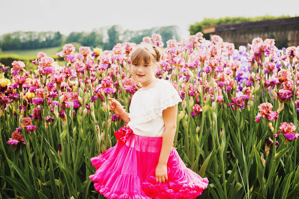 niedliches kleines Mädchen spielt im Irisblumengarten an einem schönen Sommertag, trägt weißes Hemd und leuchtend rosa Tutu-Rock - Foto, Bild