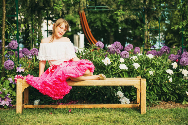 Hauska pieni tyttö lepää penkillä kauniissa kukkapuutarhassa kauniina kesäpäivänä, yllään valkoinen paita ja kirkas vaaleanpunainen tutu hame.
 - Valokuva, kuva