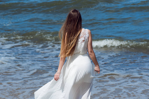 Young charming brunette woman on sea coast. Beautiful girl in a white summer dress. Runs towards the sea, back view - Foto, afbeelding