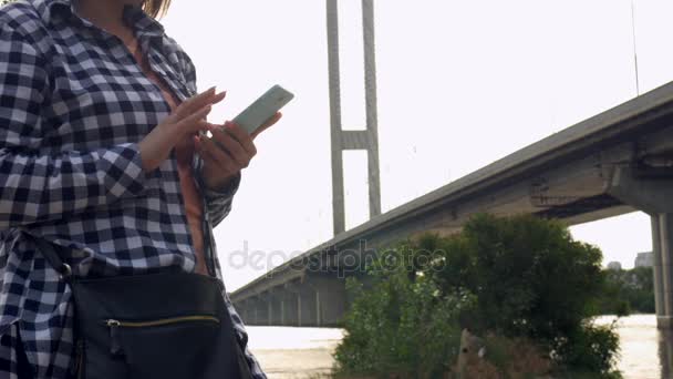 Una chica en la naturaleza está recibiendo SMS en un teléfono móvil en el fondo del puente
 - Metraje, vídeo