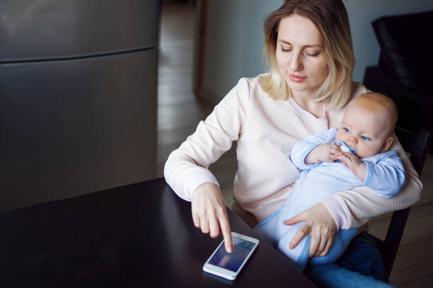 Young mother with baby sits and uses the phone. Internet browsing, indoor - Foto, Imagen