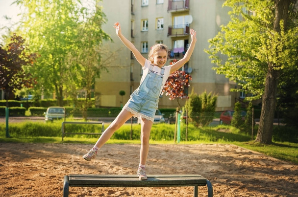 Enfant fille en salopette sautant sur un banc dans un quartier résidentiel
. - Photo, image