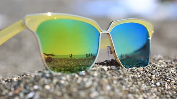 Sunglasses on the sand, the beach with the resting people is reflected in the glasses. Gold Goggles - Footage, Video