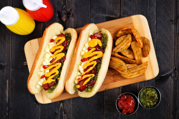 Deux hot-dogs complètement chargés avec des coins de pommes de terre sur une planche en bois
 - Photo, image
