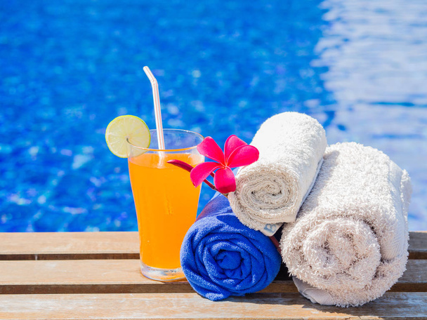 Red frangipani (plumeria) flowers, Orange juice and rolled up towels at the side of swimming pool. Vacation, beach, summer travel concept - Photo, Image