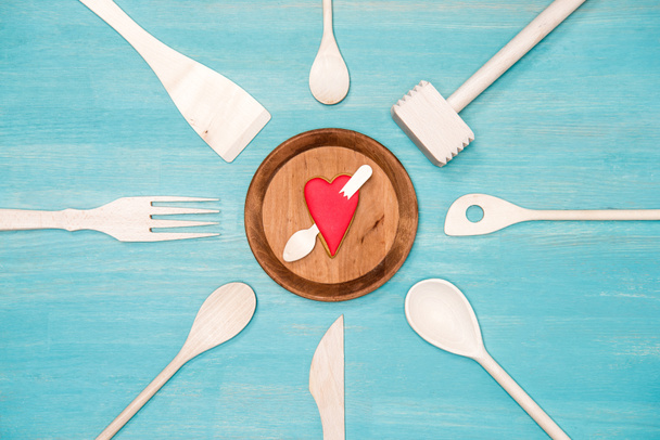 Draufsicht auf verschiedene hölzerne Küchenutensilien mit durchbohrtem Herzsymbol auf dem Teller - Foto, Bild