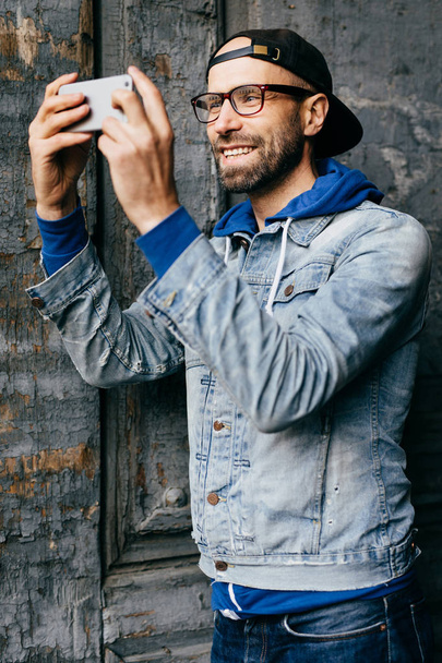 Ενθουσιασμένος αρσενικό με γένια και μουστάκι που φοράει μοντέρνα τζιν πουκάμισο με γάντζο και τζιν αναζητούν ευτυχώς κάμερα του του smartphone κάνει selfie. Εξαιρετικό hipster άνθρωπος χρησιμοποιώντας σύγχρονη συσκευή απομονωμένες - Φωτογραφία, εικόνα