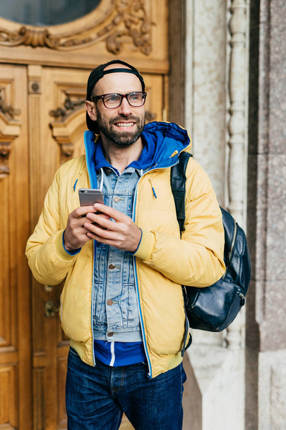 Bearded traveler with charming smile wearing trendy clothes and holding backpack standing indoors in gallery looking into distance messaging with relatives, reading sms. Stylish man having holidays - Photo, Image